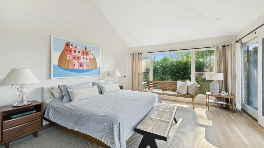 A bright bedroom with a large bed, bedside tables, and windows overlooking greenery.