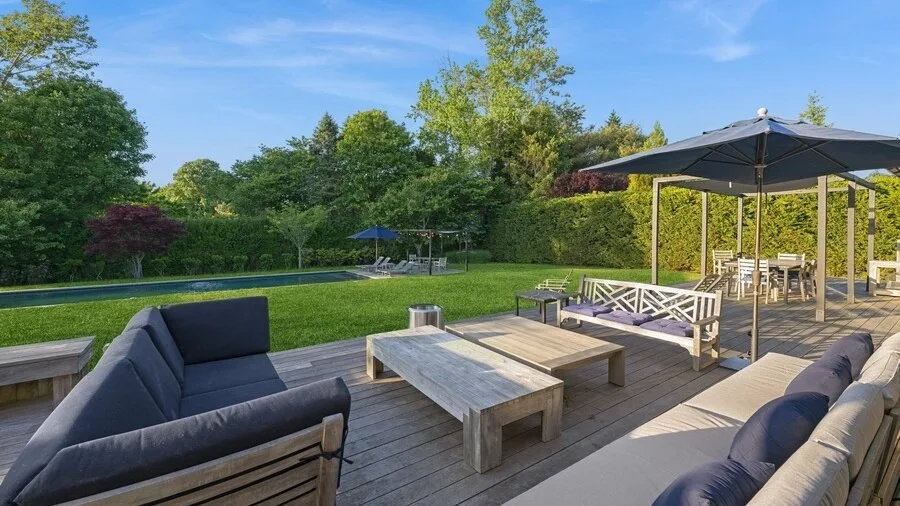 An outdoor deck with seating and a table overlooks a garden and swimming pool.