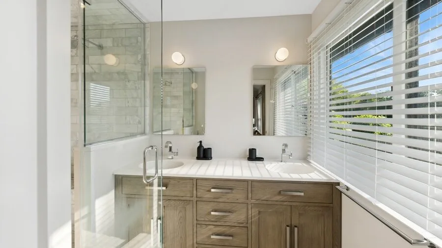 A bright bathroom with a glass shower, vanity, and window blinds.