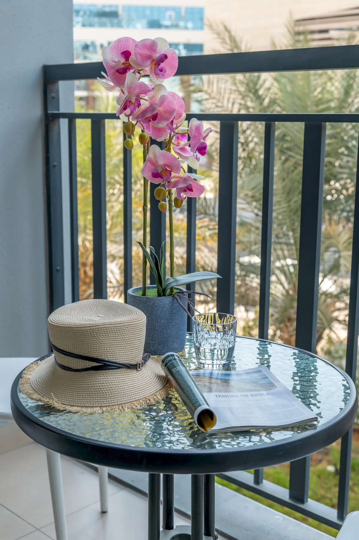 Una mesa de balcón tiene una orquídea en maceta, un sombrero y un vaso de agua.