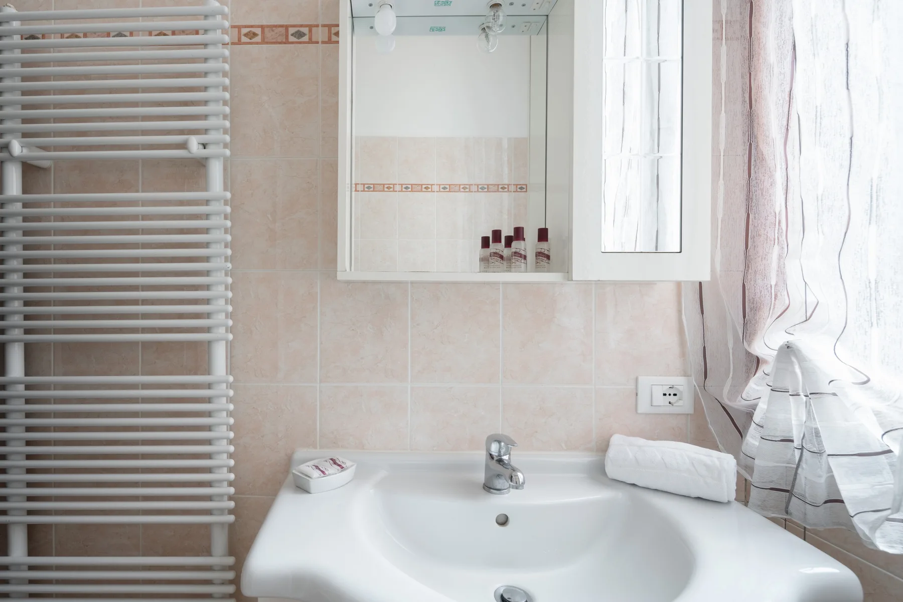 Un lavabo de baño está bajo un armario con espejo, junto a un radiador toallero y una ventana con cortinas.