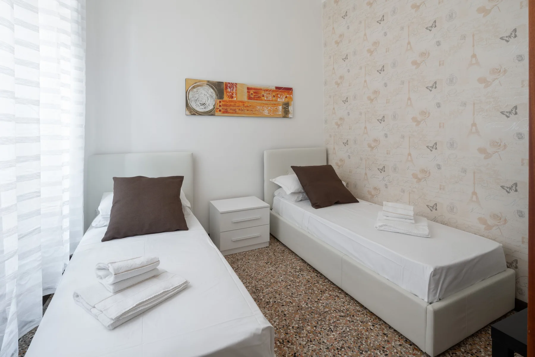 Un dormitorio luminoso con dos camas, muebles junto a ellas y papel tapiz estampado.