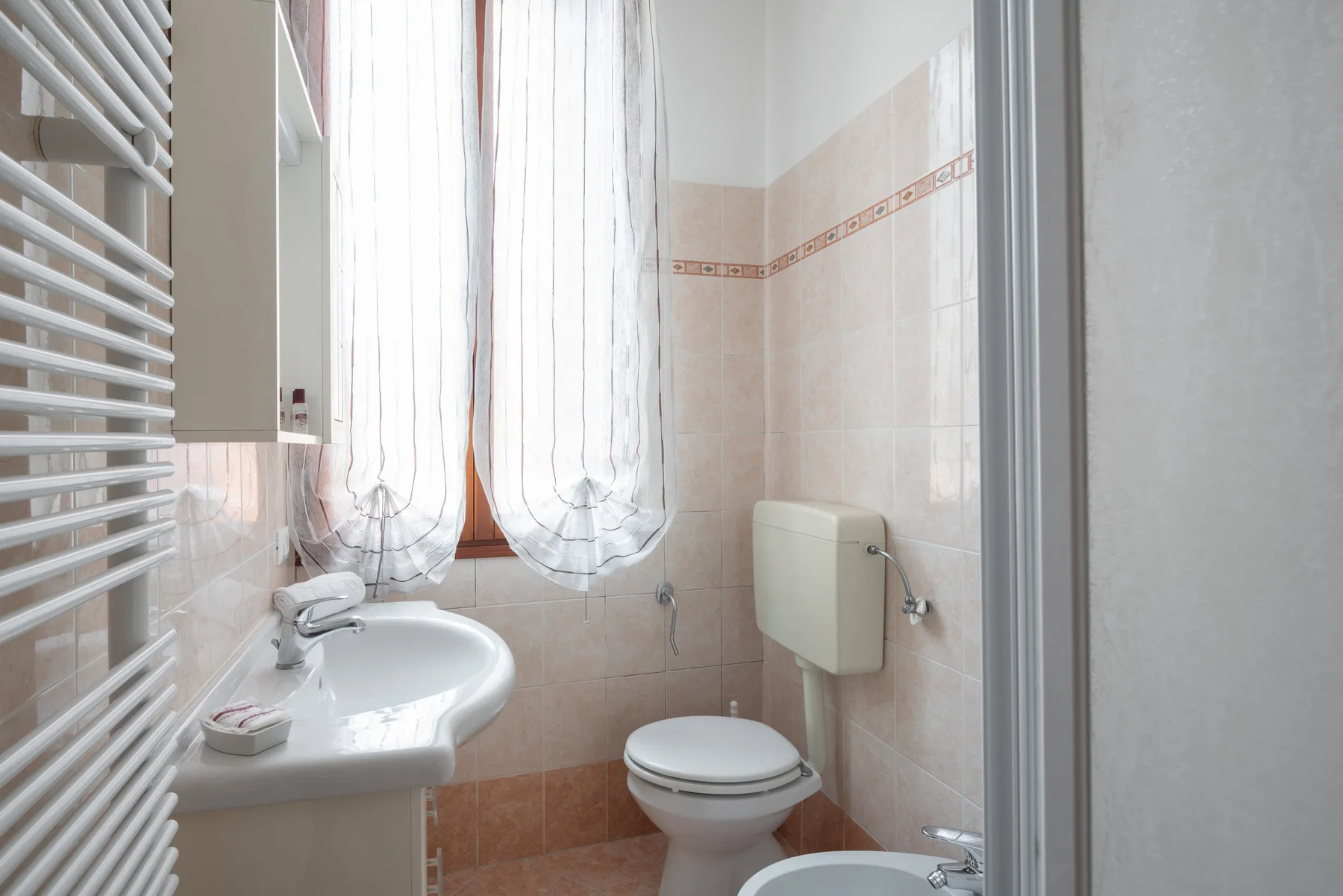 Un baño alicatado con lavabo, inodoro y una ventana cubierta con cortinas translúcidas.