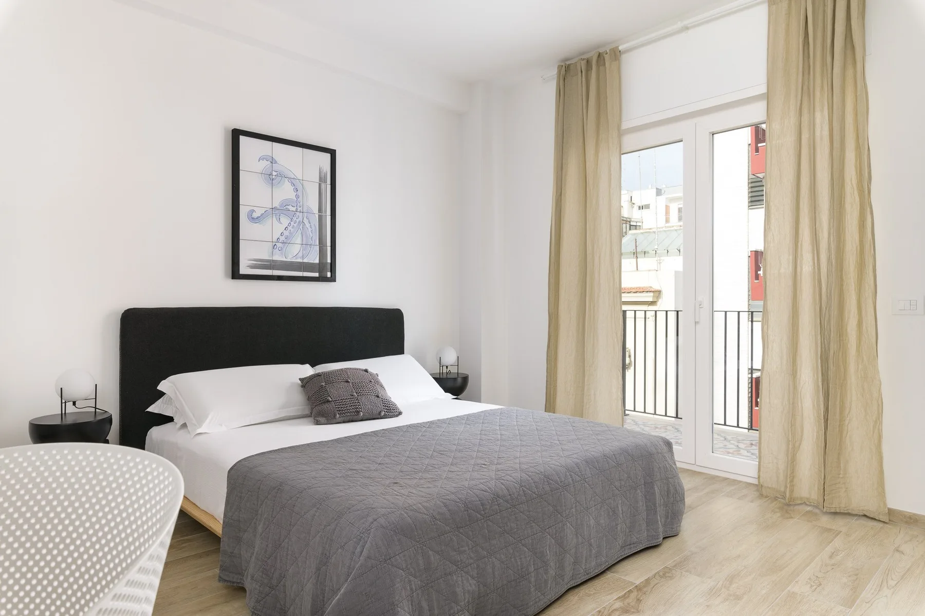 Un dormitorio luminoso con una cama junto a ventanales y cortinas beige.