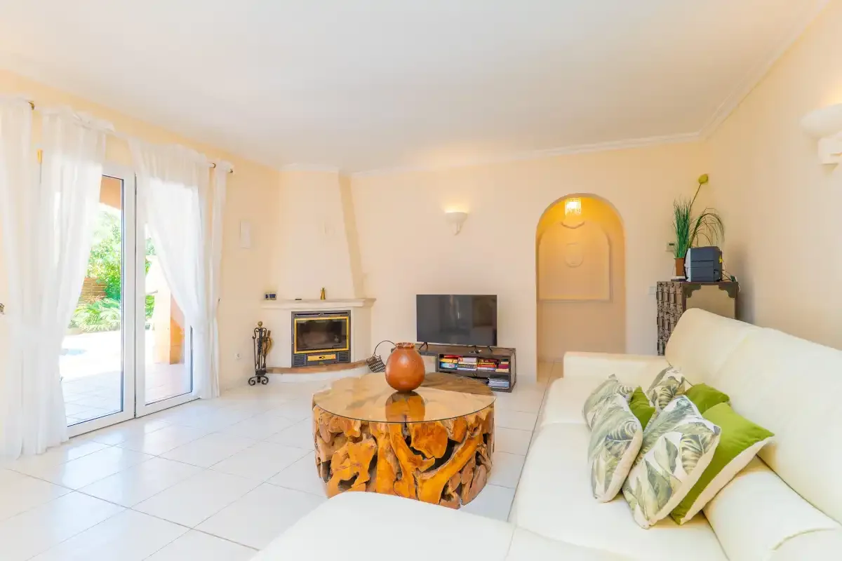 A bright living room with white seating, a television, and glass doors opening outside.