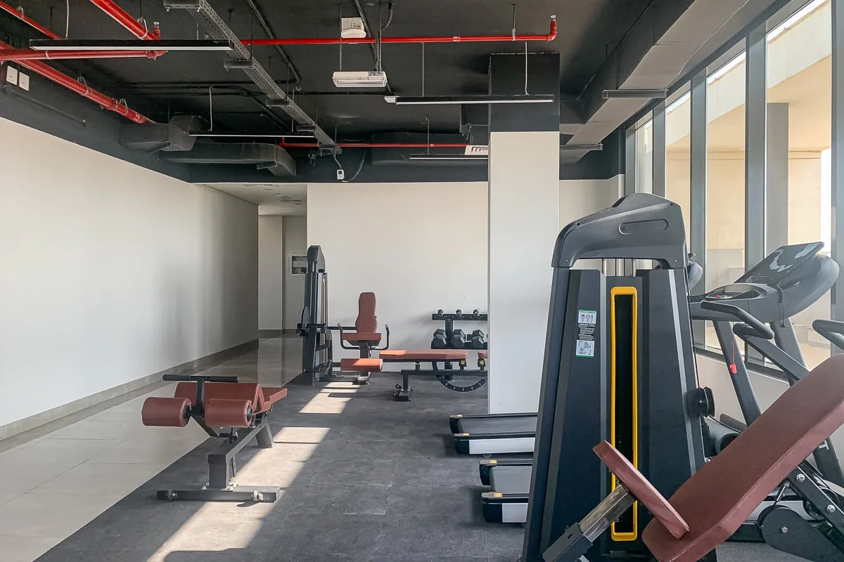 Un gimnasio con máquinas de ejercicio, pesas y grandes ventanas a lo largo de una pared.