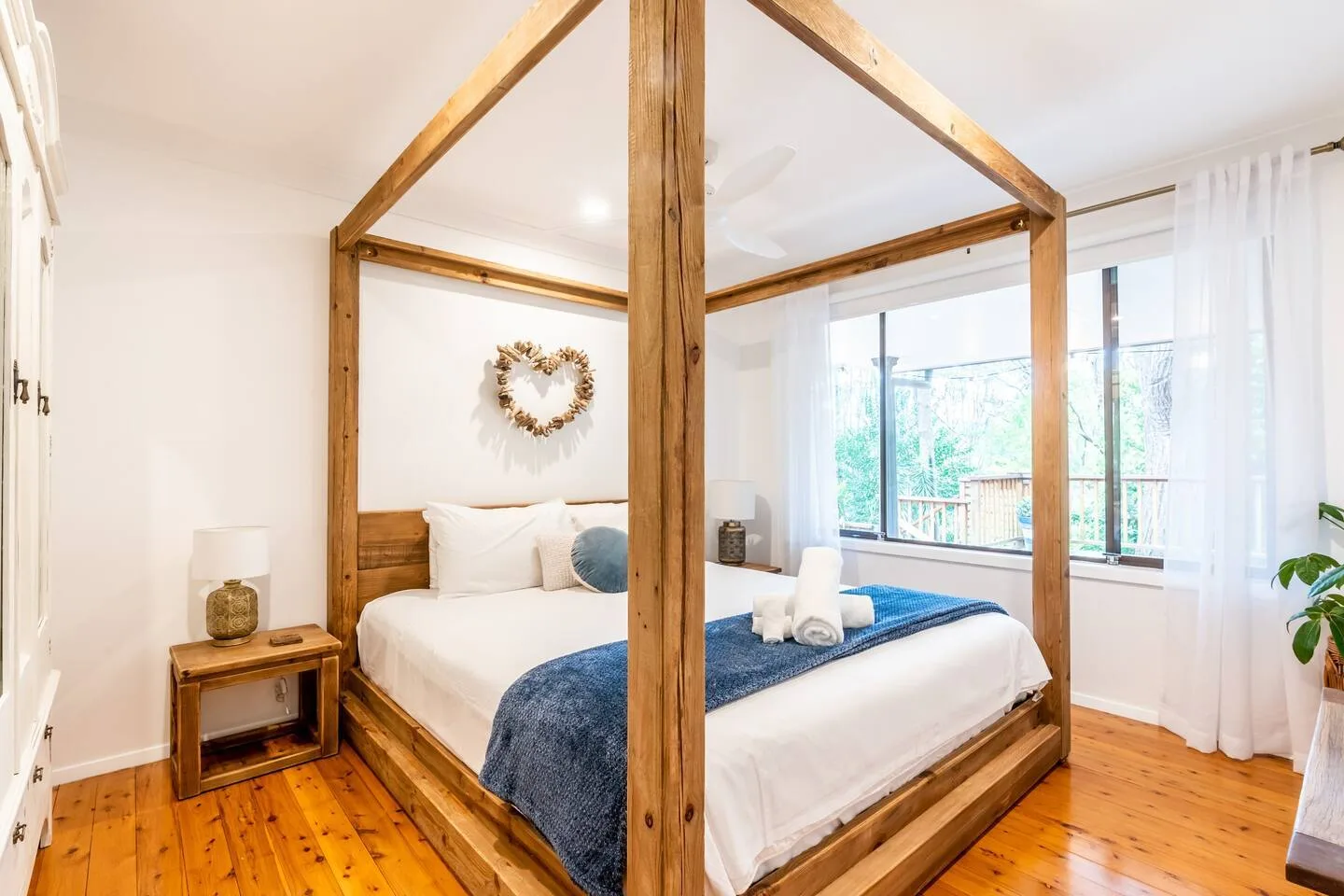 Un dormitorio luminoso tiene una cama con dosel de madera junto a grandes ventanas.