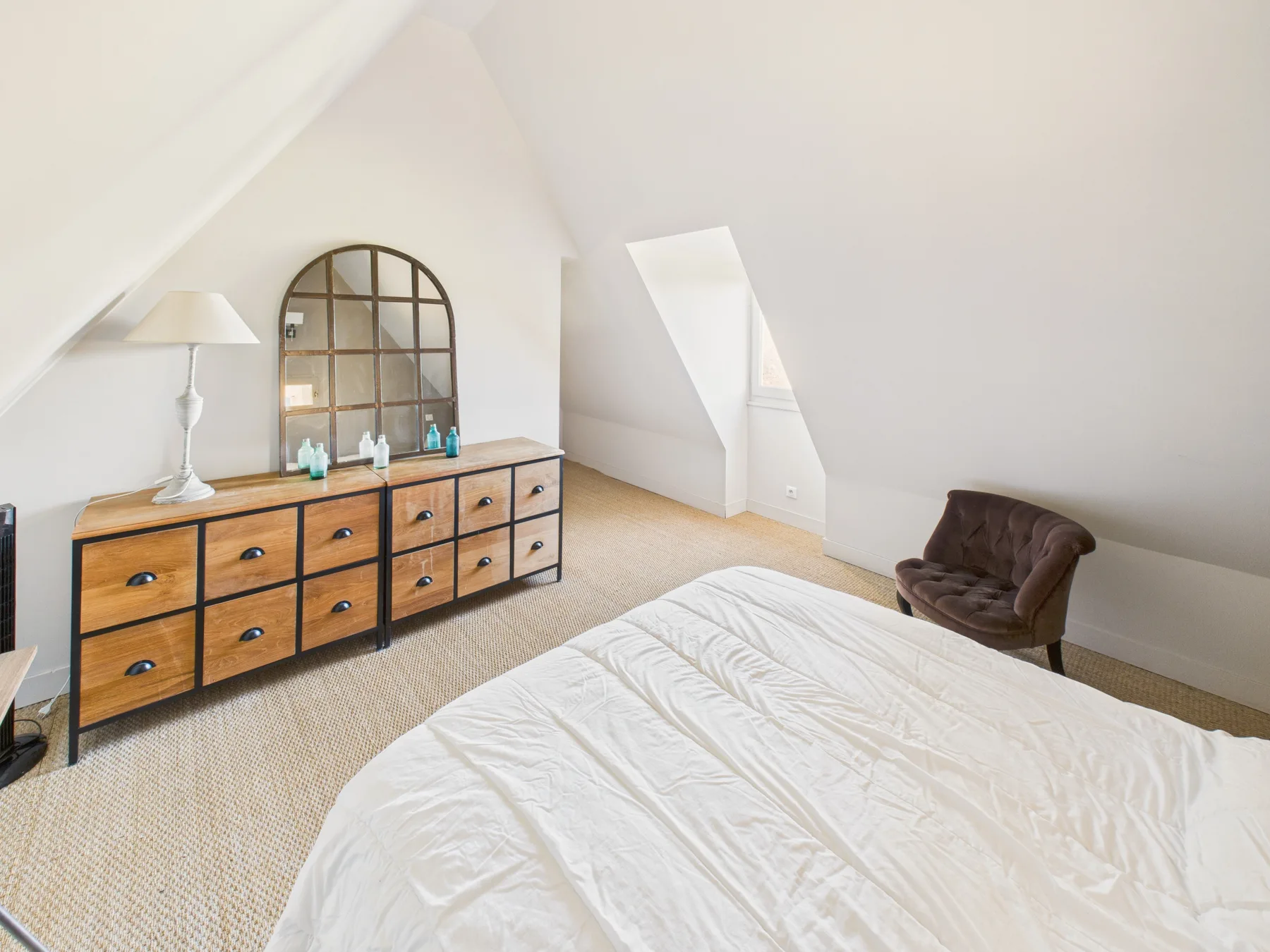 A bright attic bedroom has a bed beside a long wooden dresser.