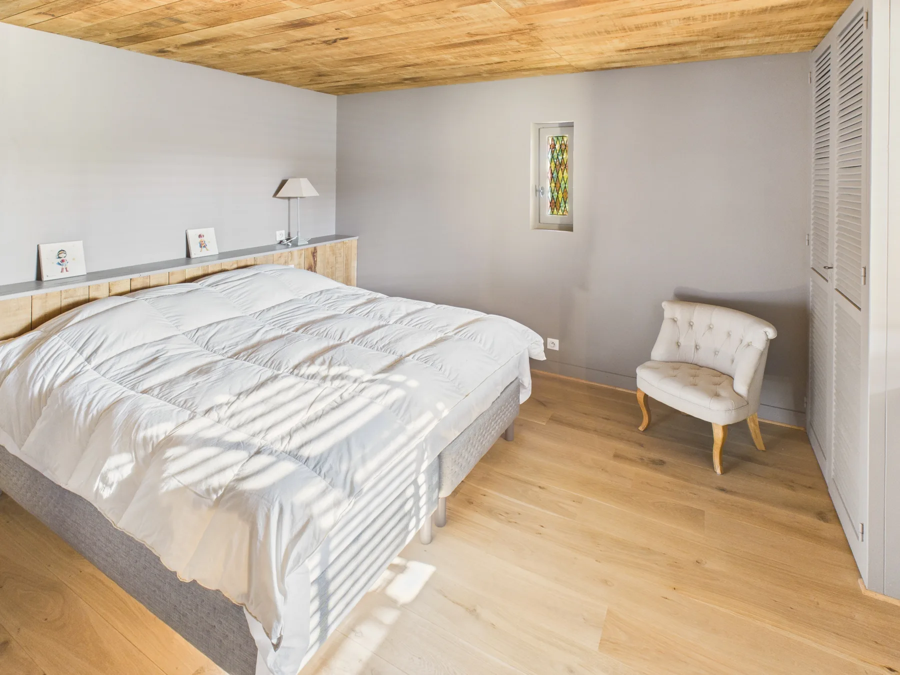 A bedroom with a bed, light wood flooring, and a small upholstered seat.