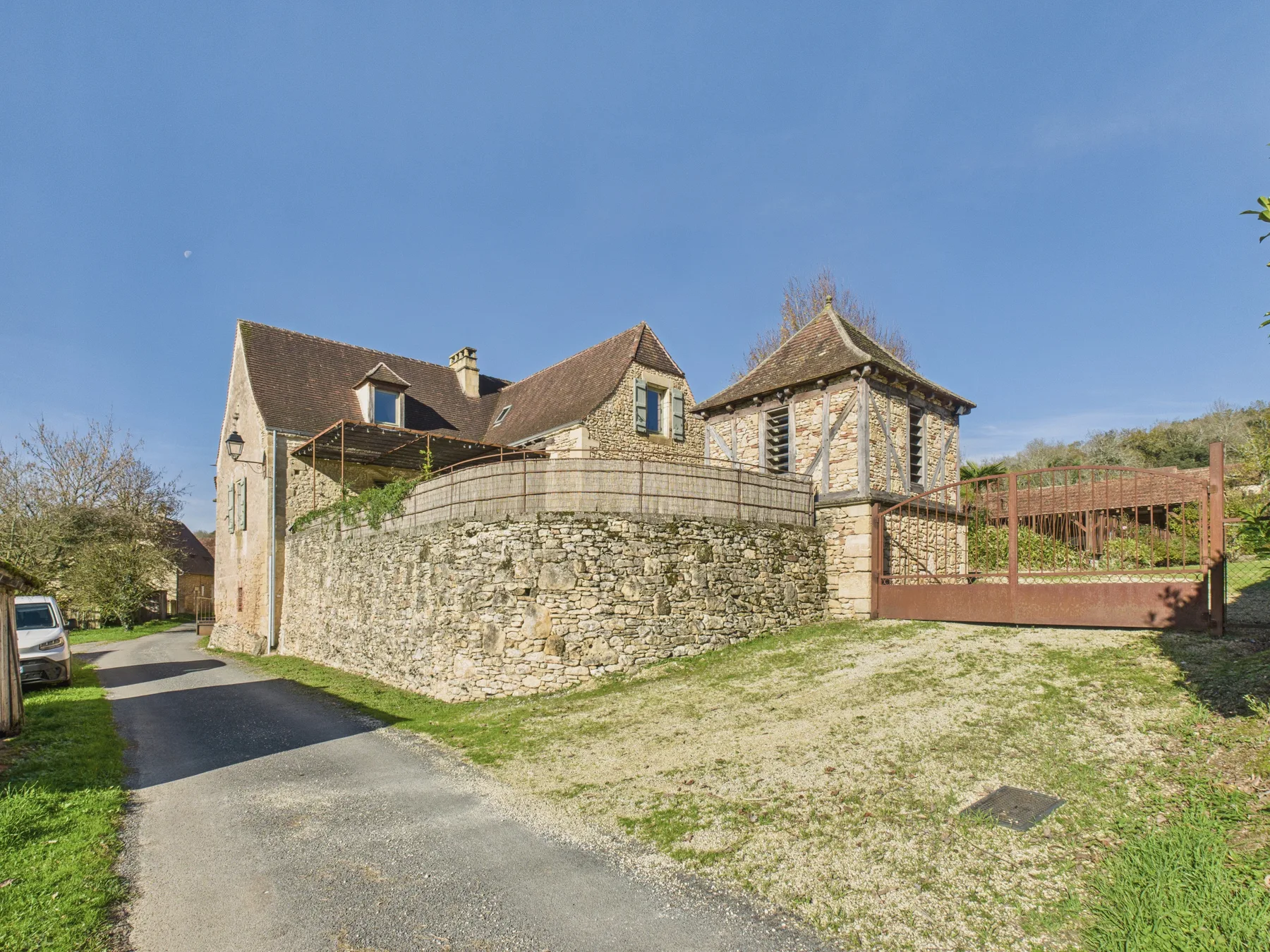 A stone house sits beside a paved driveway, with a low stone wall and wooden gate.