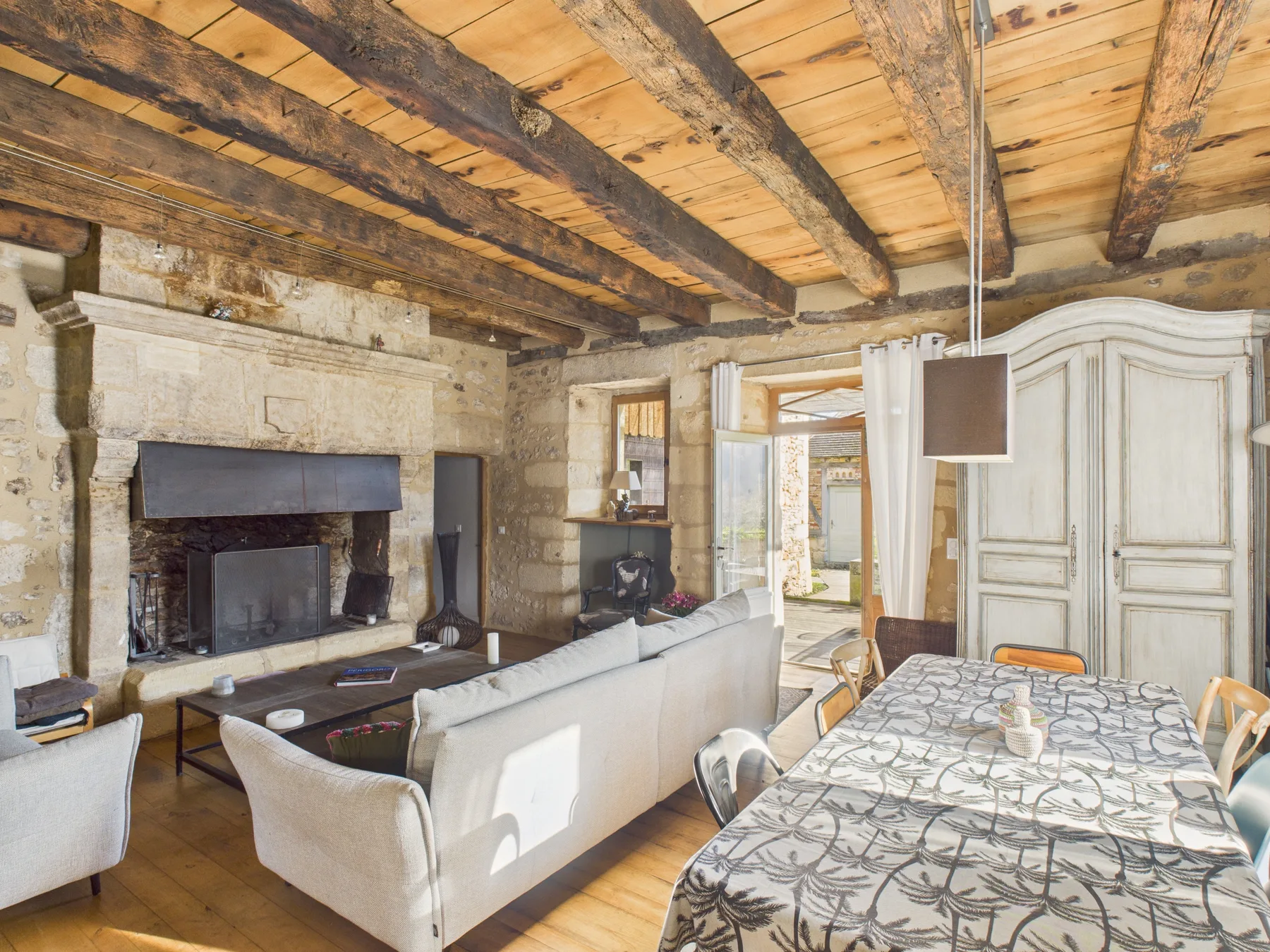 Rustic living and dining room with a large stone fireplace and exposed timber ceiling.