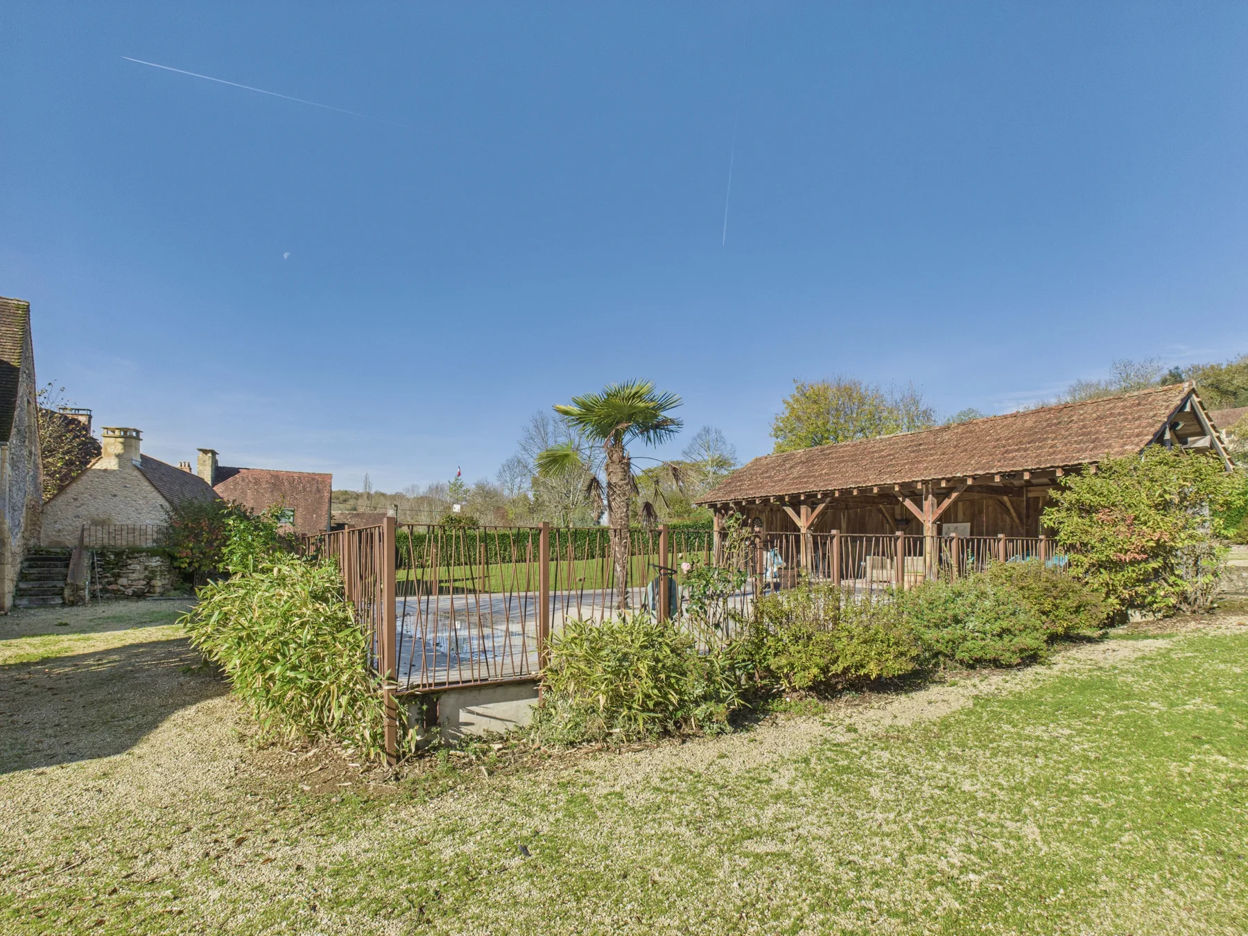 A fenced outdoor pool sits beside a covered wooden patio in a grassy yard.