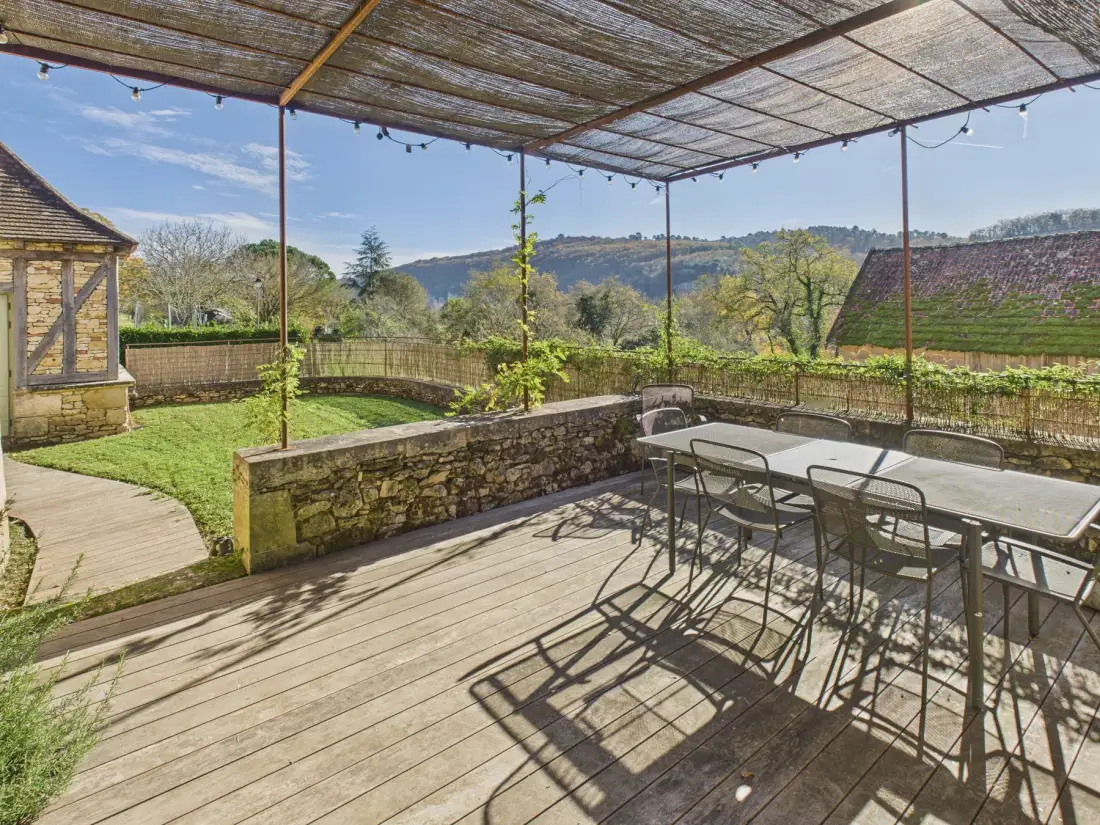 A covered outdoor dining area with a table and chairs overlooks a garden and wooded hills.