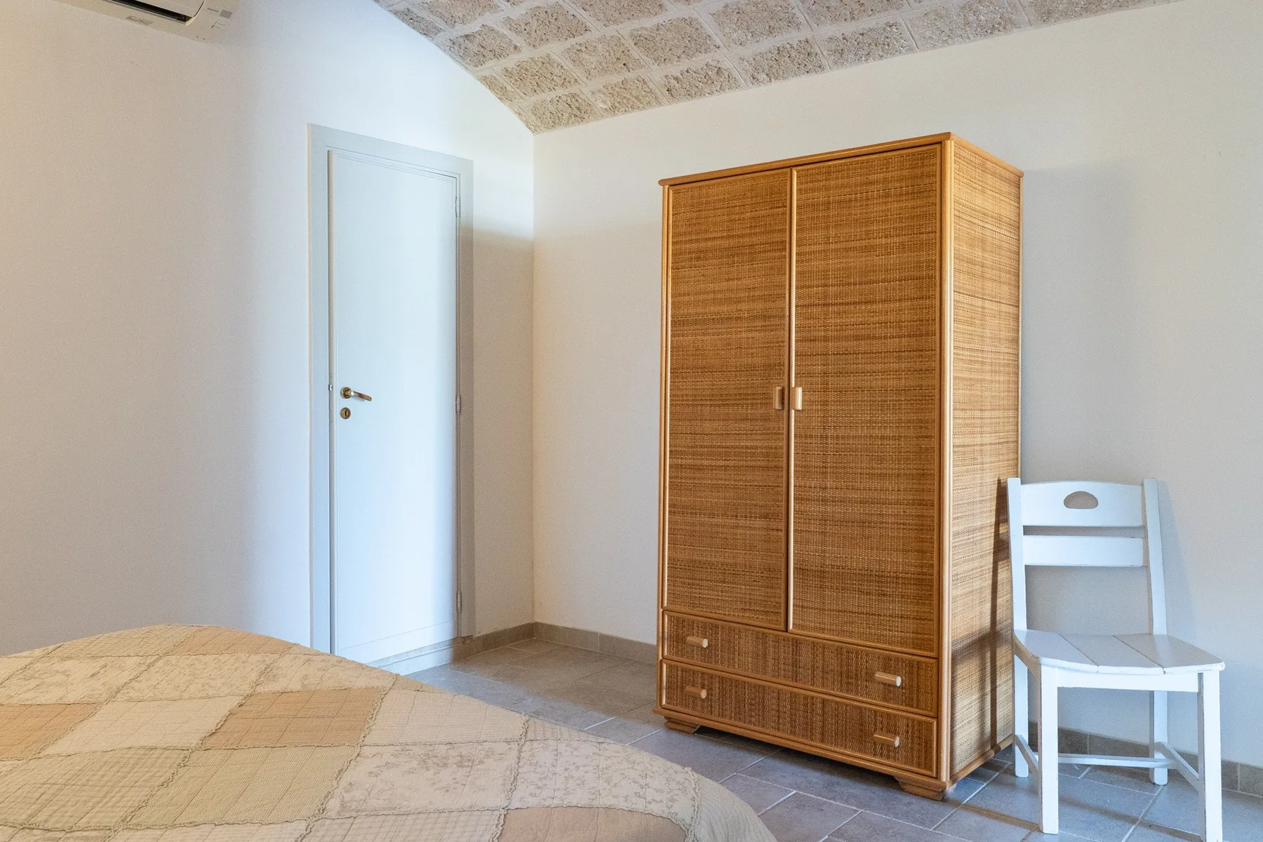 A bedroom with a bed, a tall woven-front wardrobe, and a white chair.
