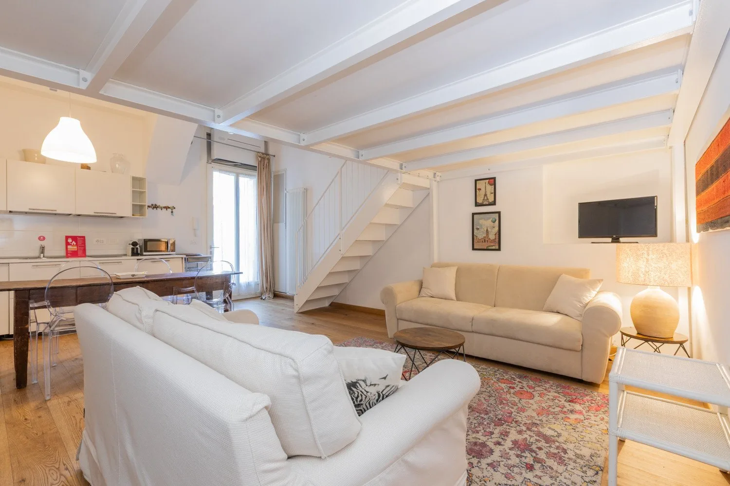 A bright living room with seating, a rug, and a staircase beside the kitchen.