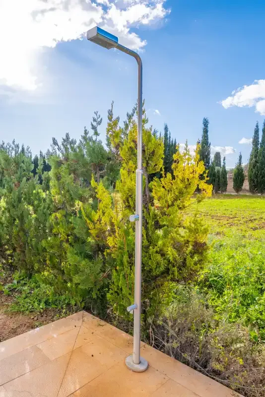 An outdoor shower stands on a tiled patio beside green shrubs.