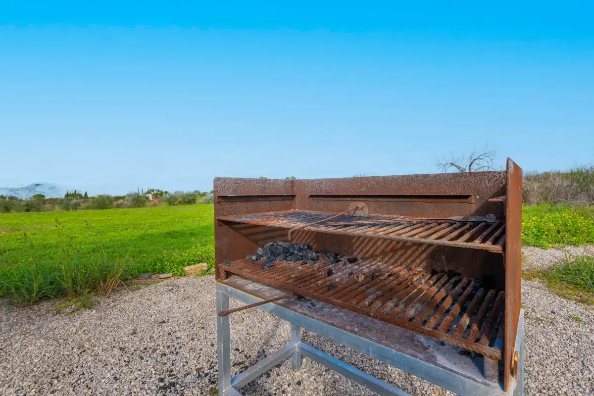 A metal barbecue grill stands on a gravel area beside a green field.