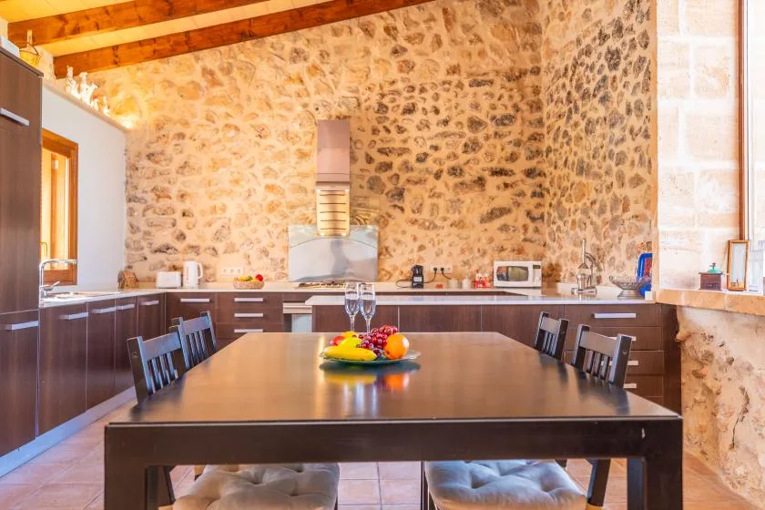 A dining table sits in a kitchen with stone walls and wooden cabinets.