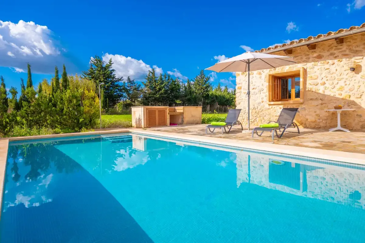 A swimming pool beside a stone building, with loungers and an umbrella on the patio.