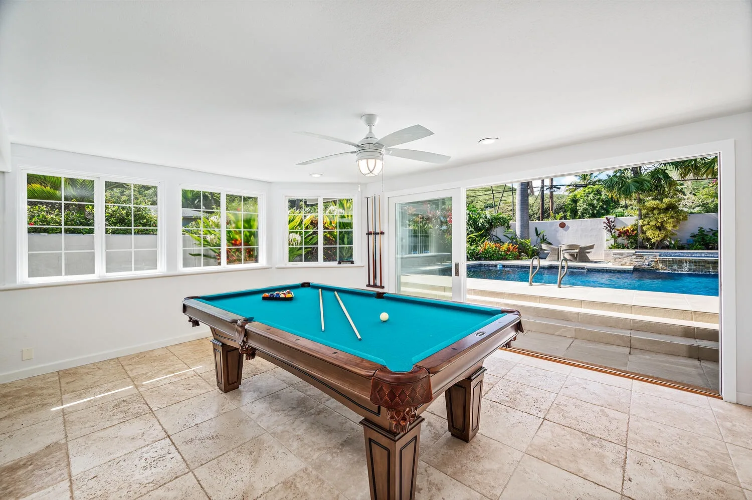 Una mesa de billar está en una habitación luminosa junto a ventanas con vista a una piscina.