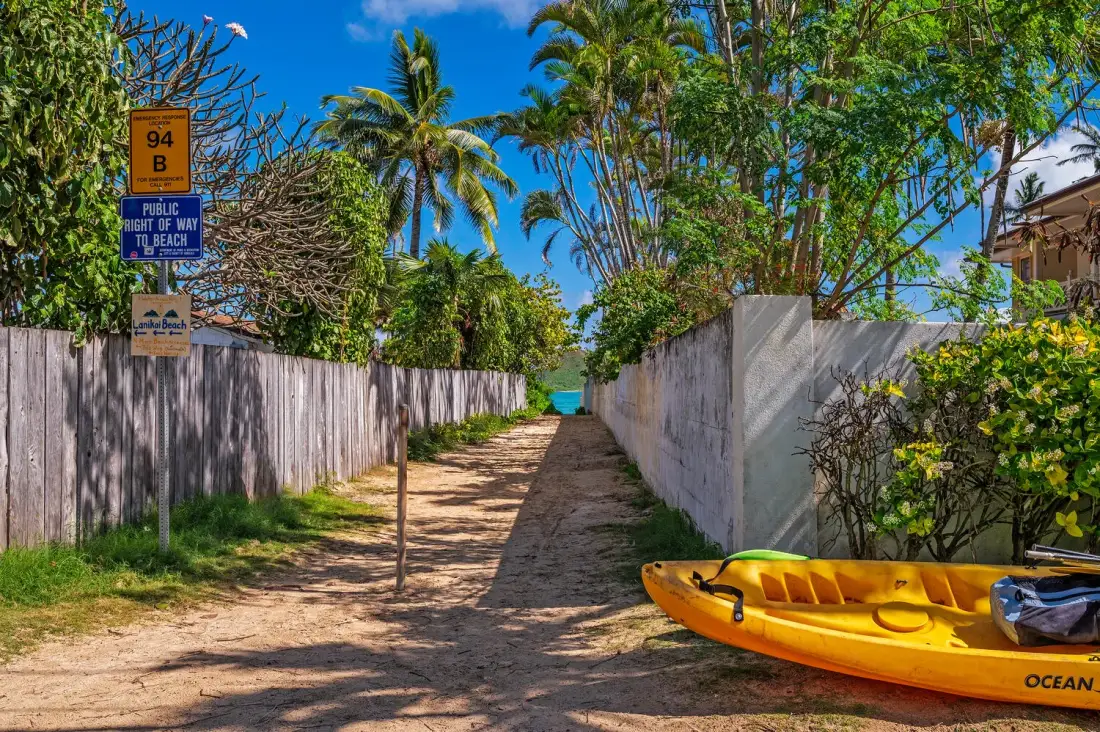 Un sendero de arena entre cercas conduce hacia el agua, con un kayak amarillo al lado.
