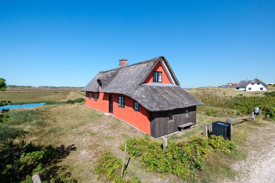 A red thatched cottage stands among open grassy fields under a clear blue sky.