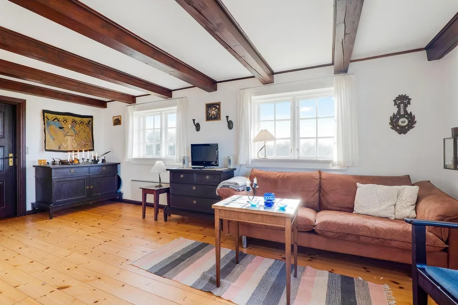 A bright living room with a brown sofa, wooden furniture, and exposed ceiling beams.