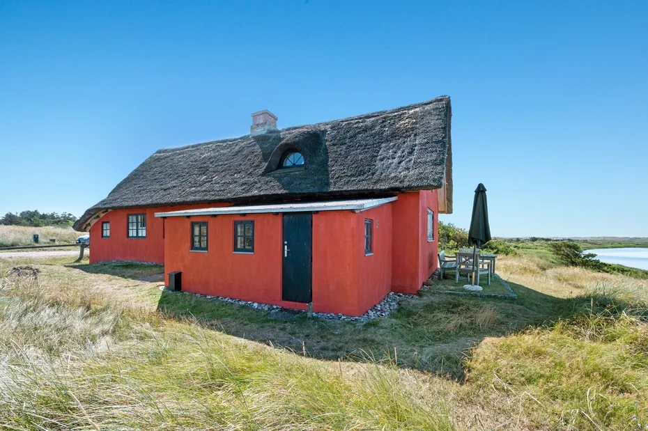 A red cottage stands in an open grassy landscape beneath a thatched roof.