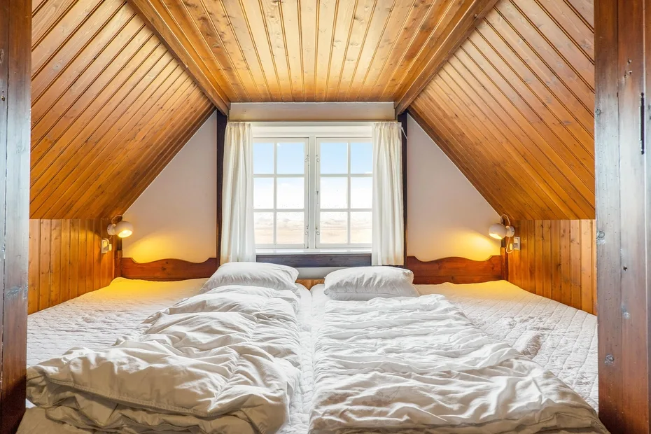 A bedroom with two beds beneath a sloped wooden ceiling and a central window.