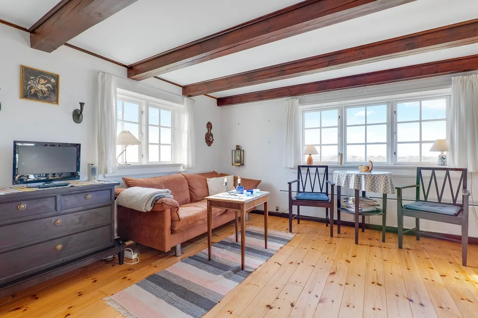 A bright living room has wooden ceiling beams, seating, and large windows.