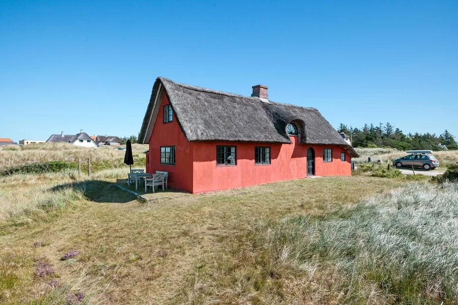 A red cottage with a thatched roof stands in an open grassy landscape.