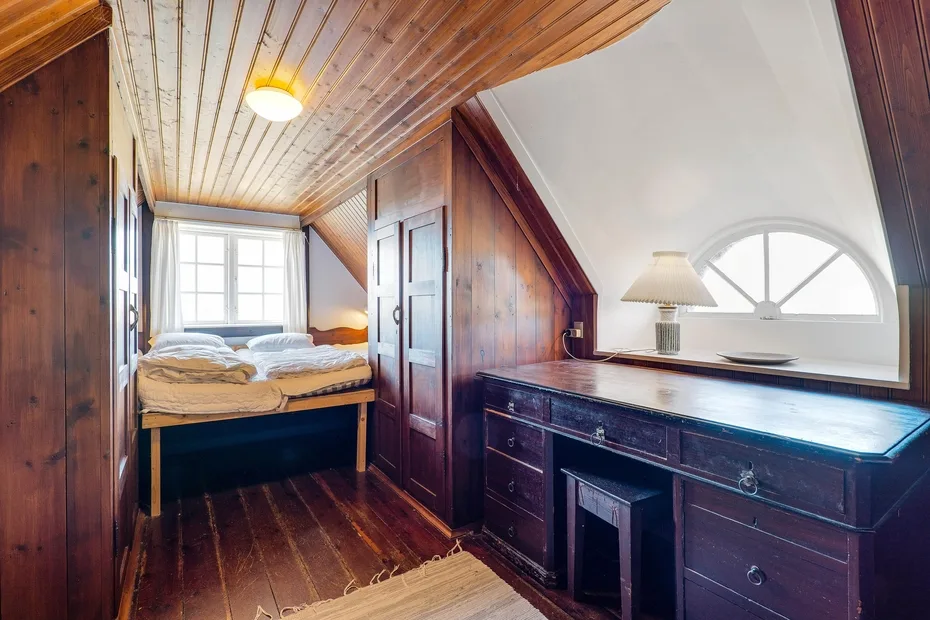 A wood-paneled bedroom with a bed and a long dresser beneath an arched window.