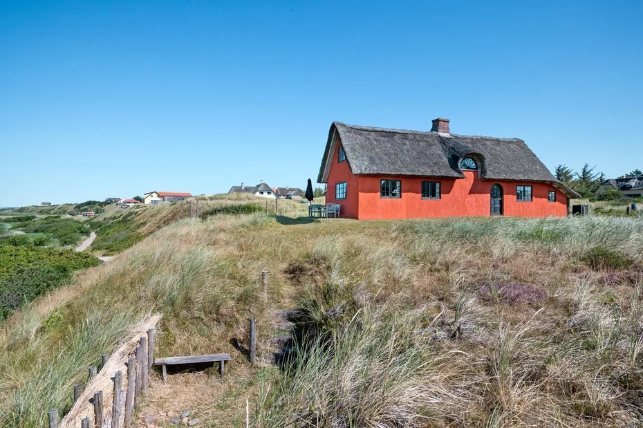 A red cottage sits among coastal grasses beneath a clear blue sky.