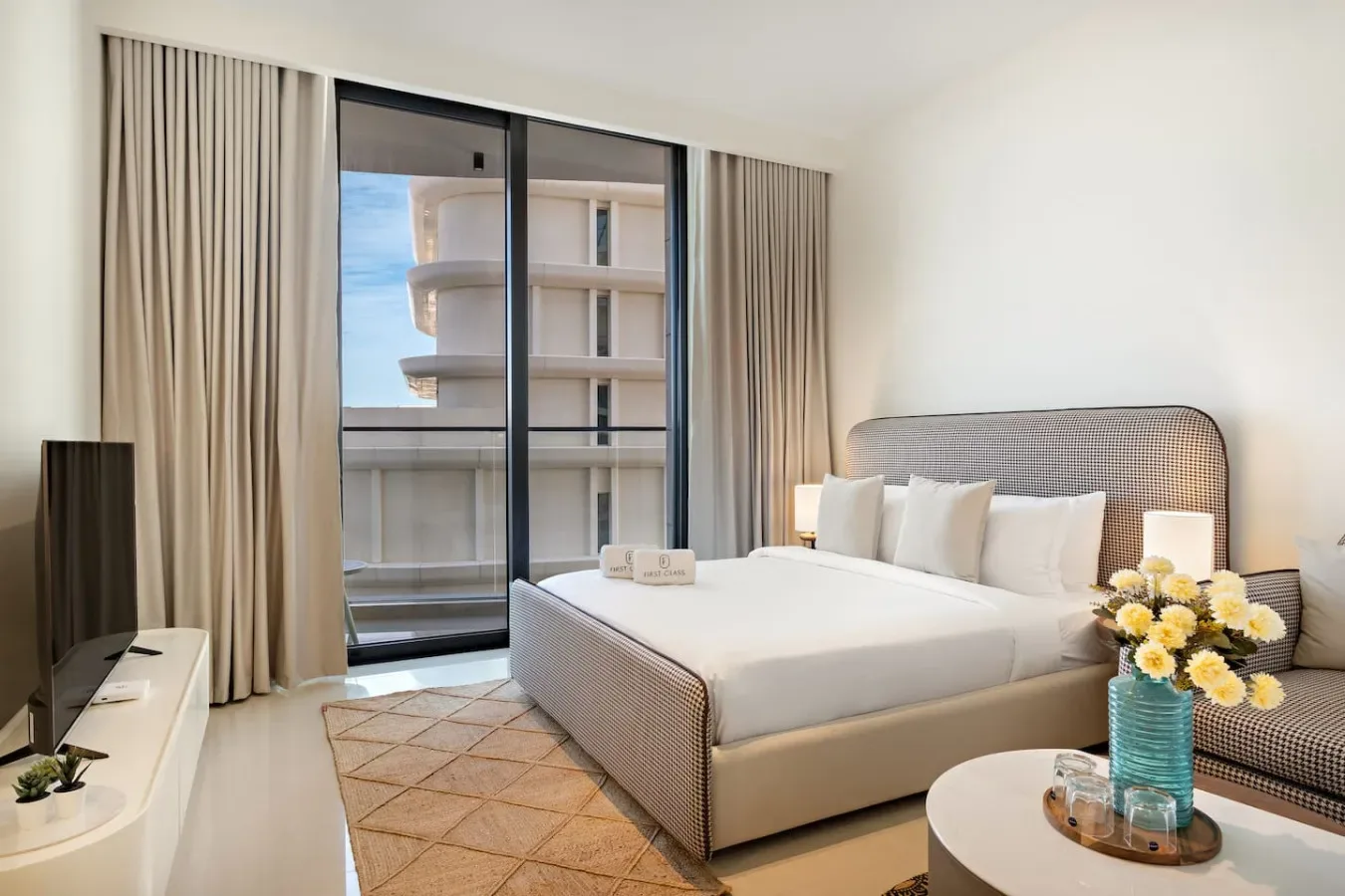 Un dormitorio luminoso con una cama grande, lámparas de noche y cortinas junto a una puerta de vidrio.