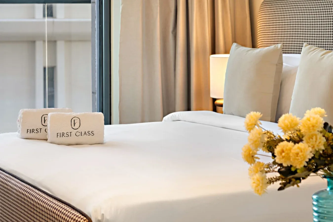 Una cama bien tendida con almohadas claras, una lámpara de noche y un pequeño arreglo floral.