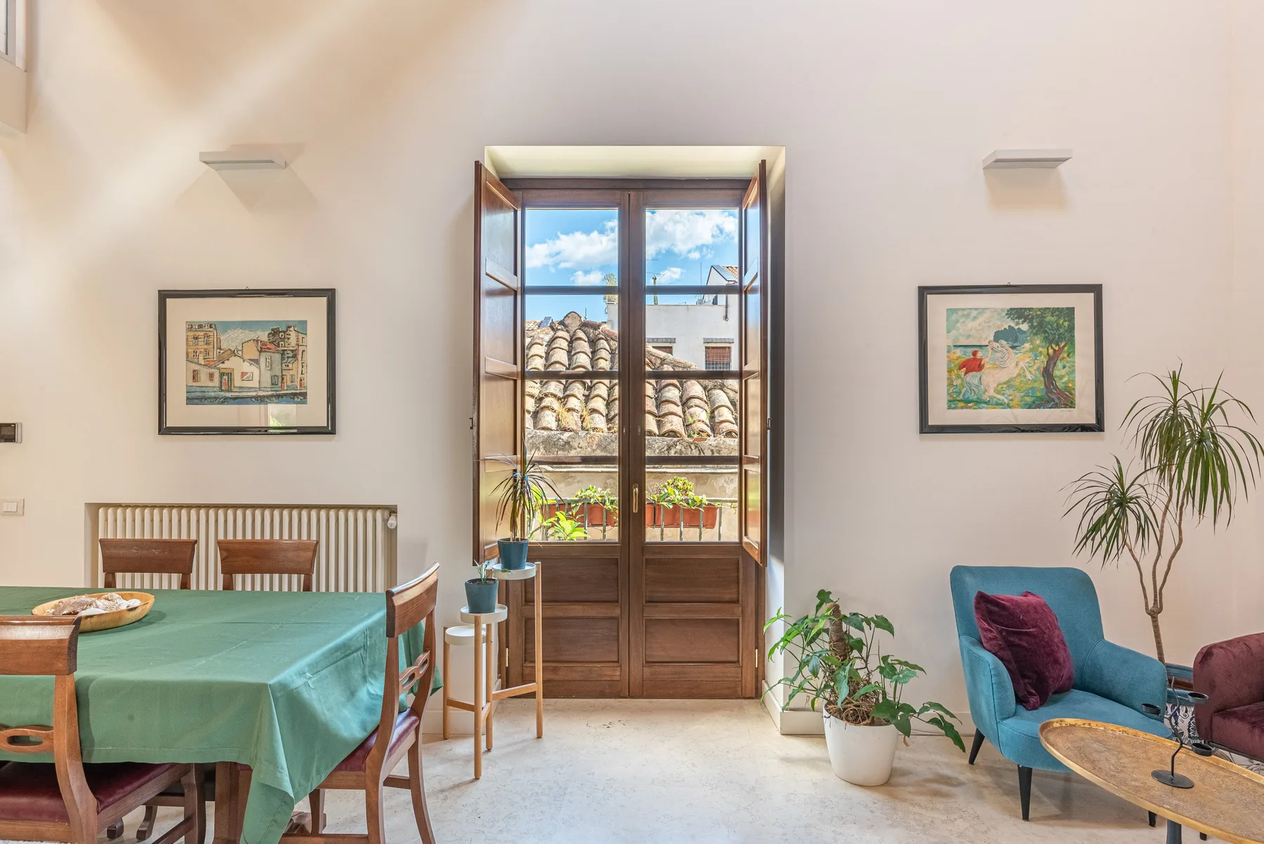 A bright dining and sitting room with a tall window overlooking nearby rooftops.