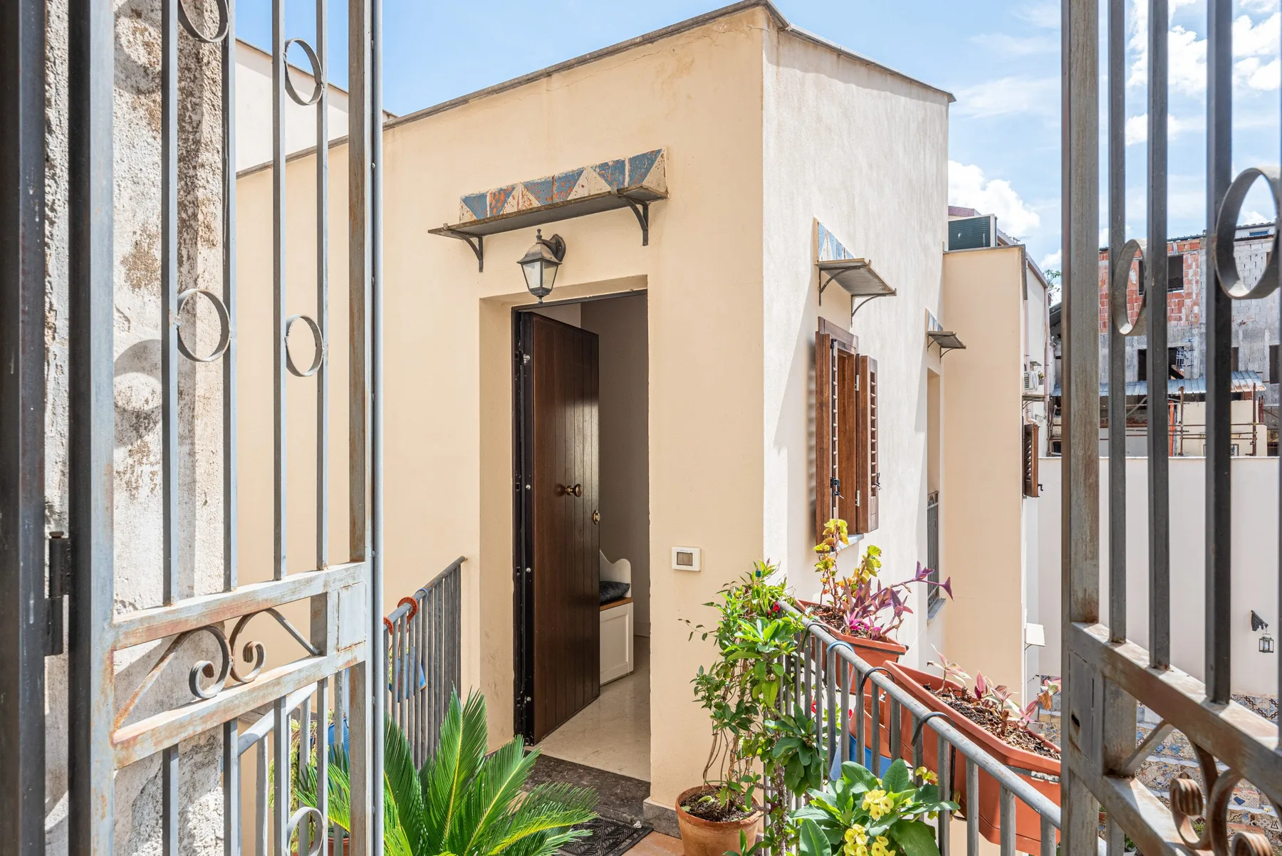 A small building entrance opens onto a sunlit walkway lined with potted plants.