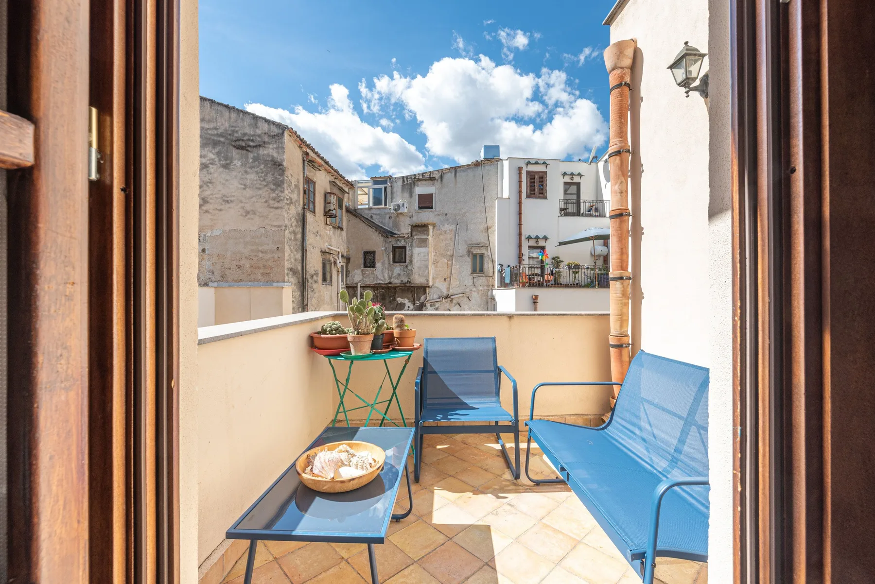A sunny terrace with blue seating and potted plants overlooks nearby buildings.