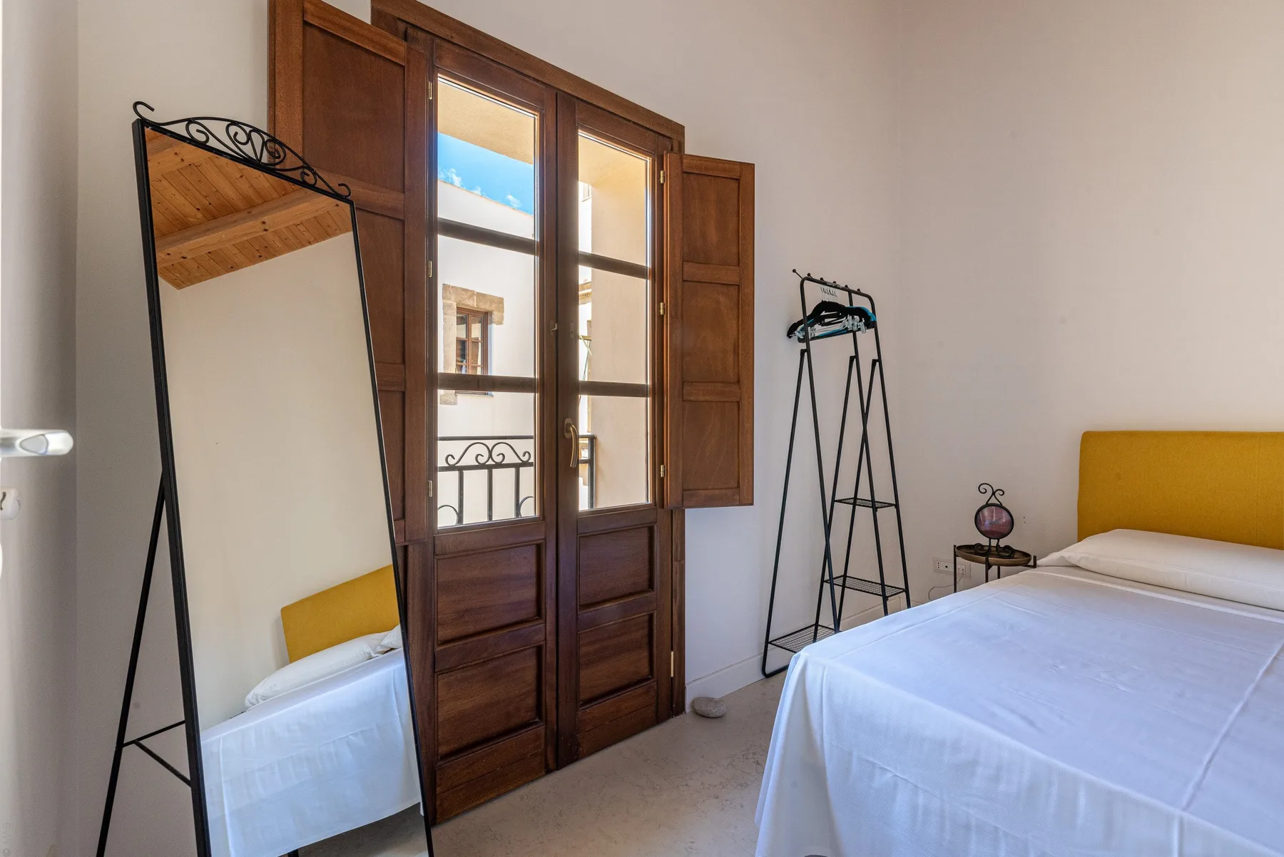 A bedroom with a bed beside tall wooden windows and a freestanding mirror.
