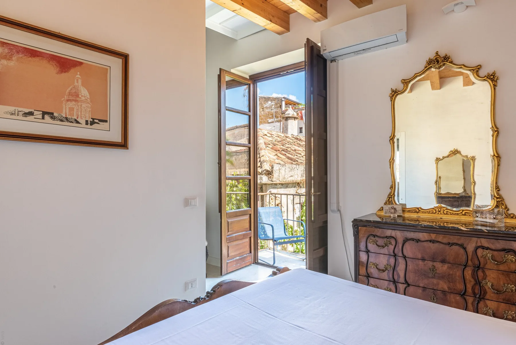 A bedroom with an open balcony door and an ornate mirror above a dresser.
