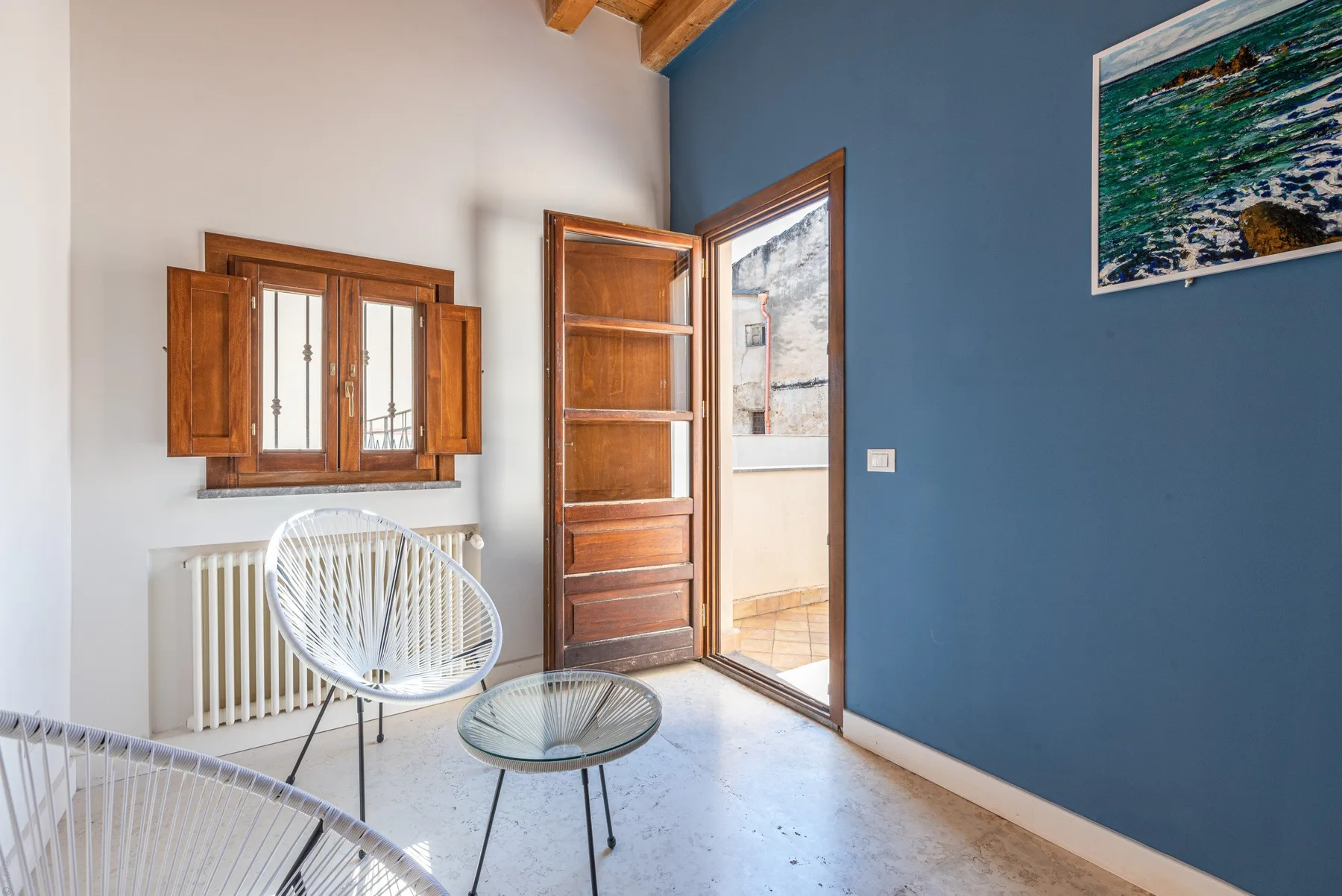 A bright room with wooden shutters, seating, and an open doorway beside a blue wall.