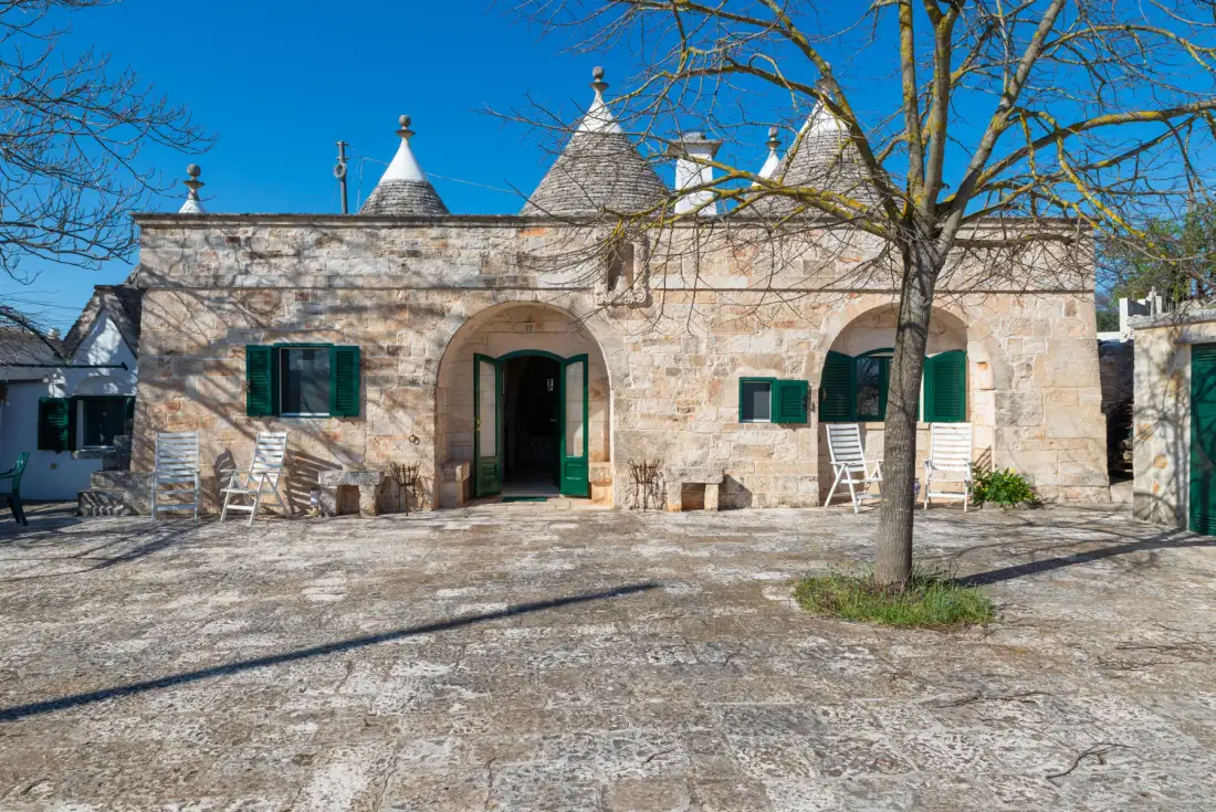 Una casa de piedra con contraventanas verdes y una puerta arqueada abierta bajo un cielo despejado.