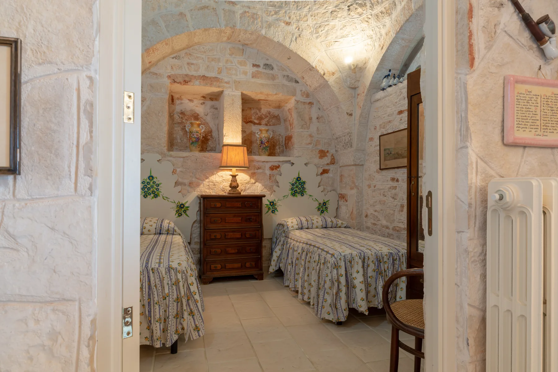 Dormitorio de piedra con dos camas y una lámpara encendida sobre una cómoda.
