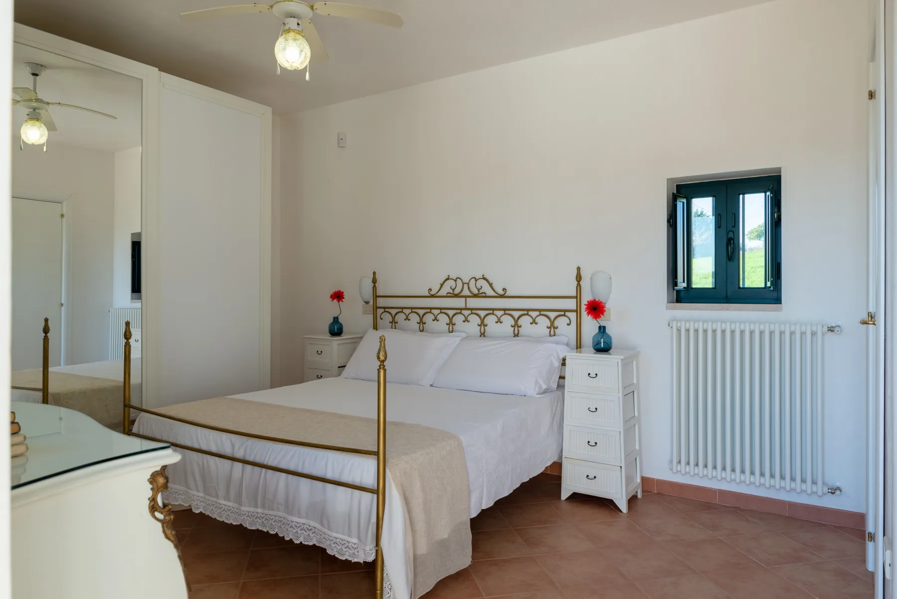 Un dormitorio con una cama de latón, ropa de cama blanca y una ventana sobre un radiador.
