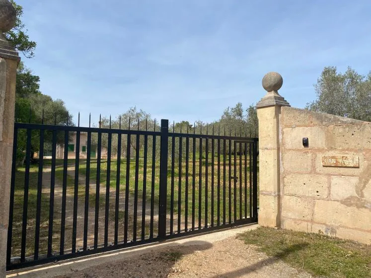 Una puerta metálica negra está junto a un muro de piedra, frente a una zona arbolada.
