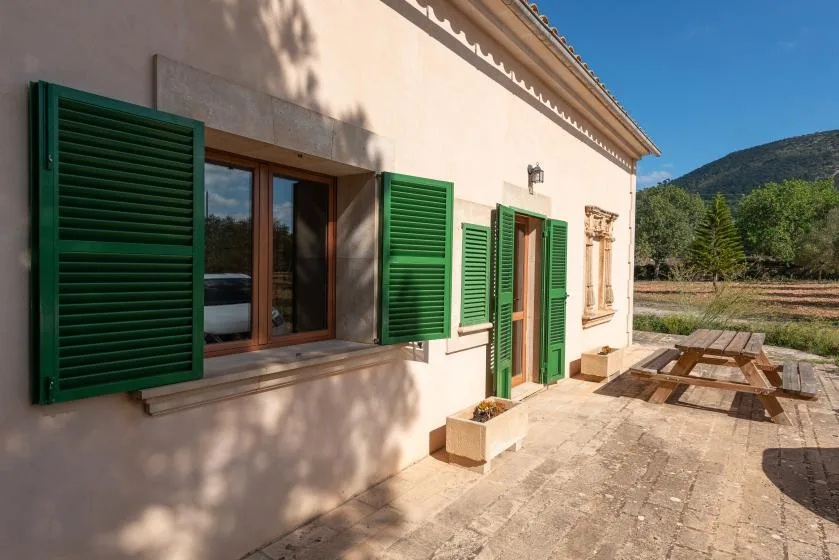 El exterior soleado de un edificio con contraventanas verdes da a una terraza de piedra con asientos de picnic.