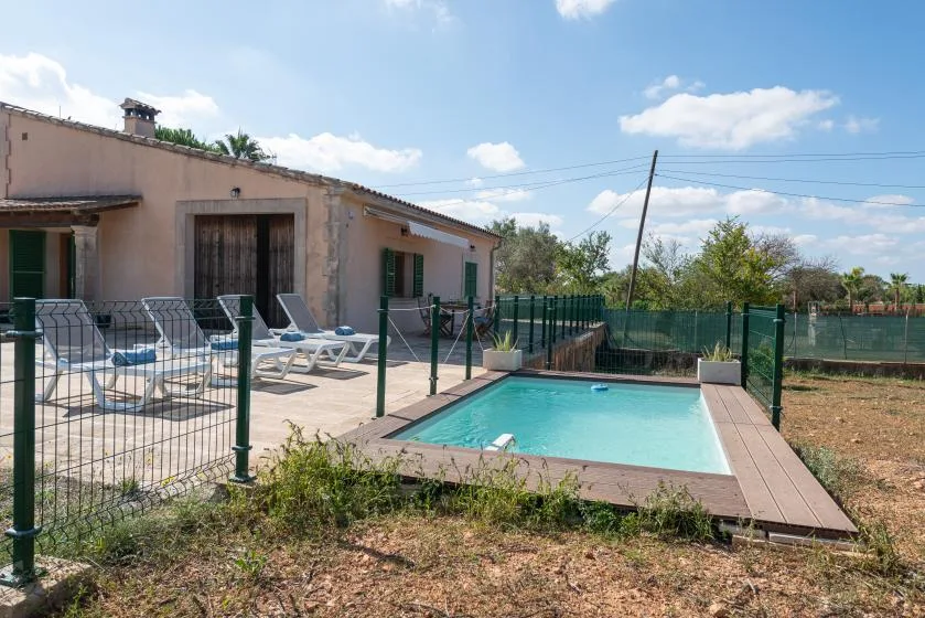 Piscina al aire libre junto a una casa de una planta, con tumbonas en el patio pavimentado.