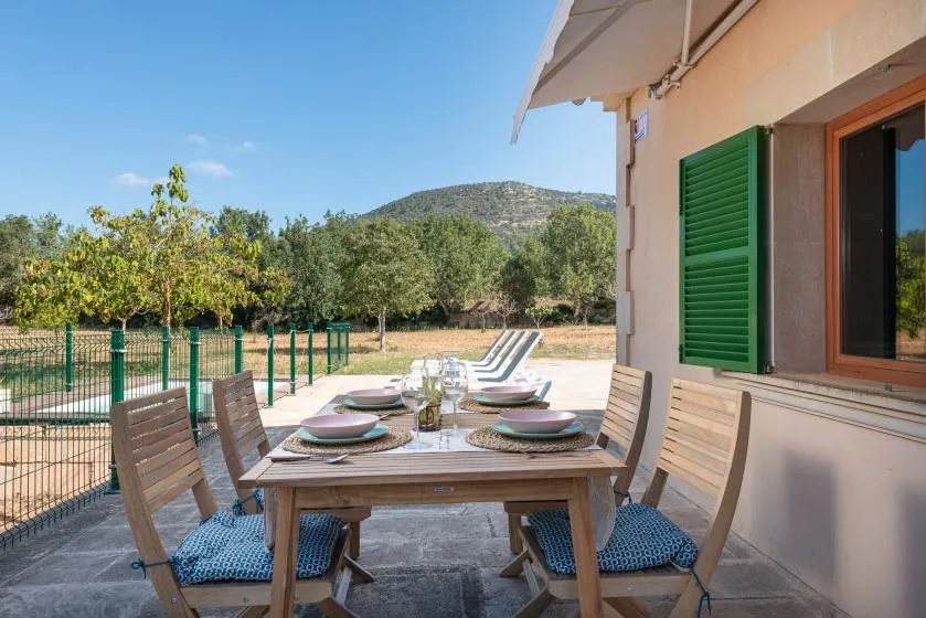 Comedor al aire libre junto a una casa, con mesa y asientos frente a un jardín.