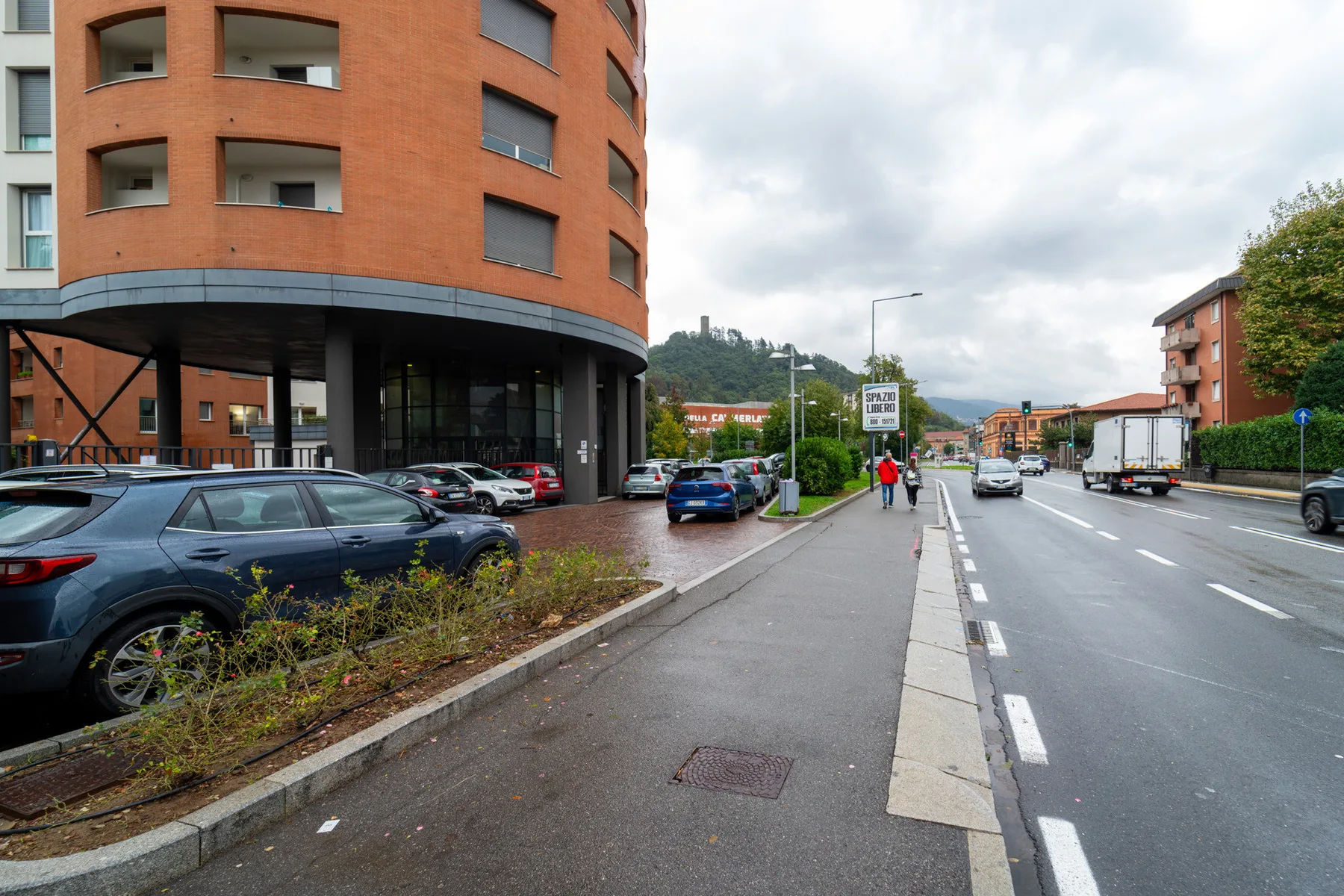 A city street beside a rounded brick building, with parked cars along the sidewalk.