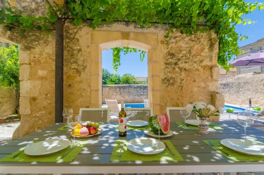 Una mesa de comedor está bajo una pérgola frondosa, junto a un arco de piedra y una piscina.